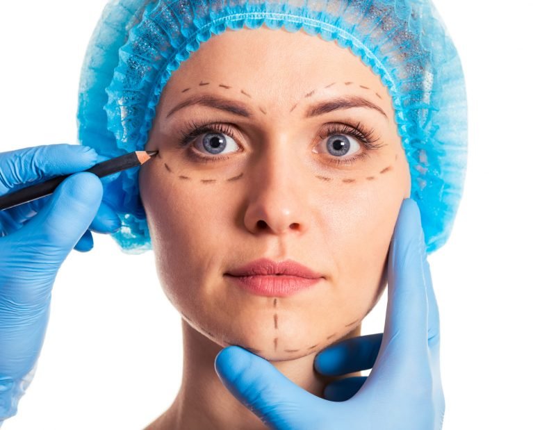 Beautiful woman in medical headwear is looking at camera, surgeon in medical gloves is making sketches on face, isolated on white background