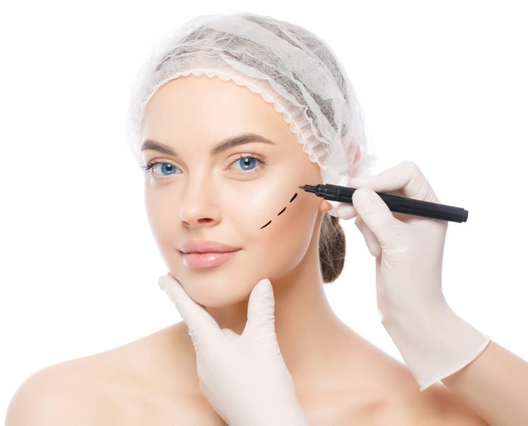 Young beautiful woman prepared for plastic surgery by doctor who uses skin marker, isolated on white background